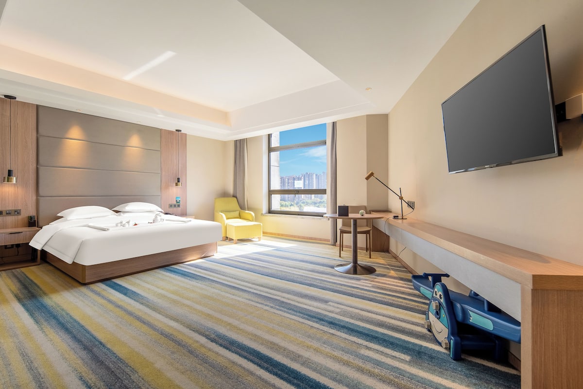A modern hotel room with a large bed, a desk, a chair, a TV, and a view of the city through the window.