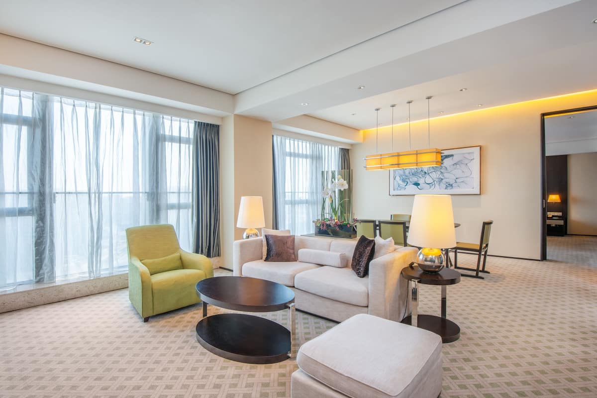 A modern hotel room with a large window, a sofa, a chair, a small table, and a lamp.