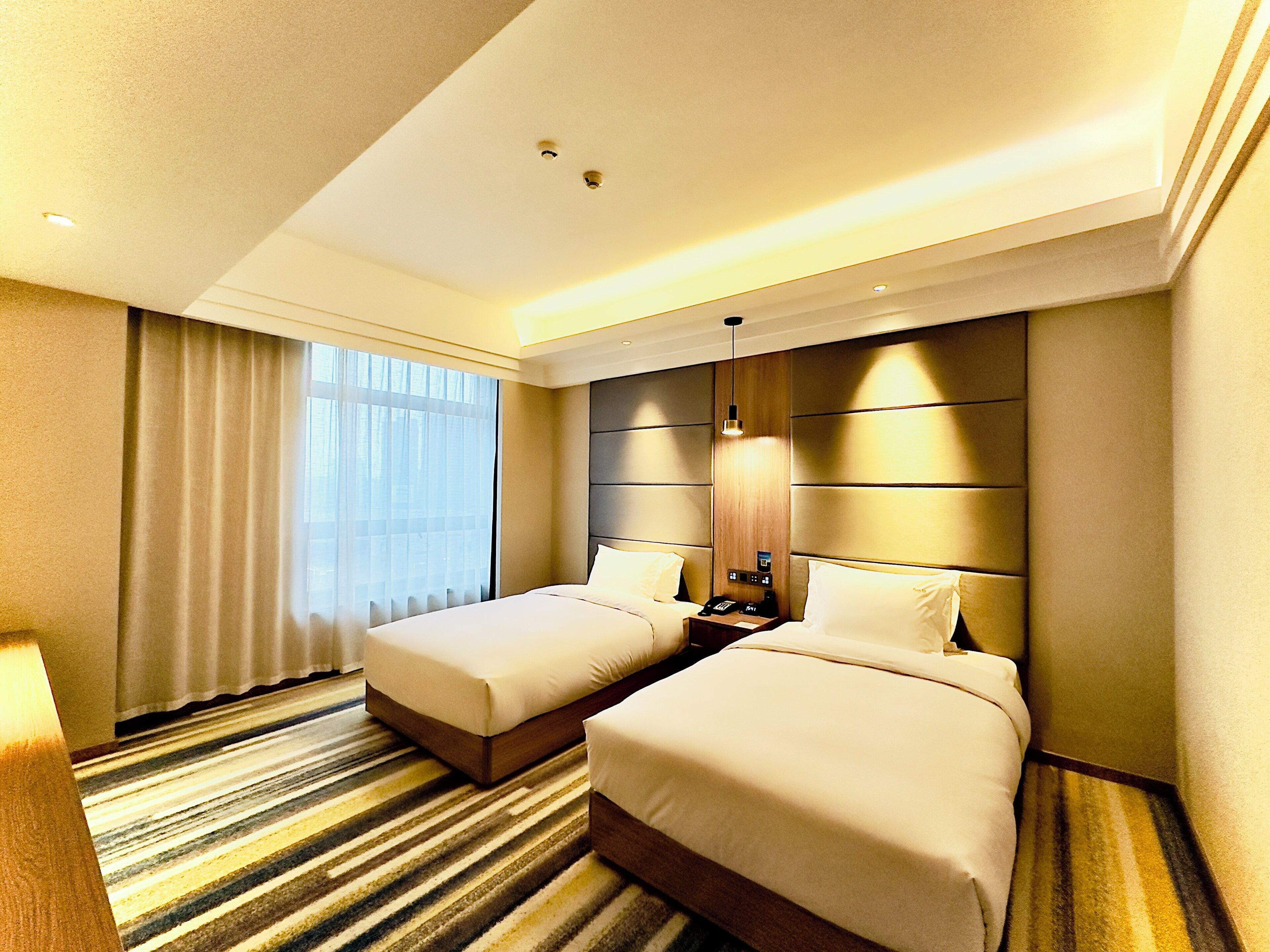 Ein Hotelzimmer mit zwei Betten, einem großen Fenster mit Vorhängen, einem Streifenmuster-Teppich und einem modernen Kopfteil.