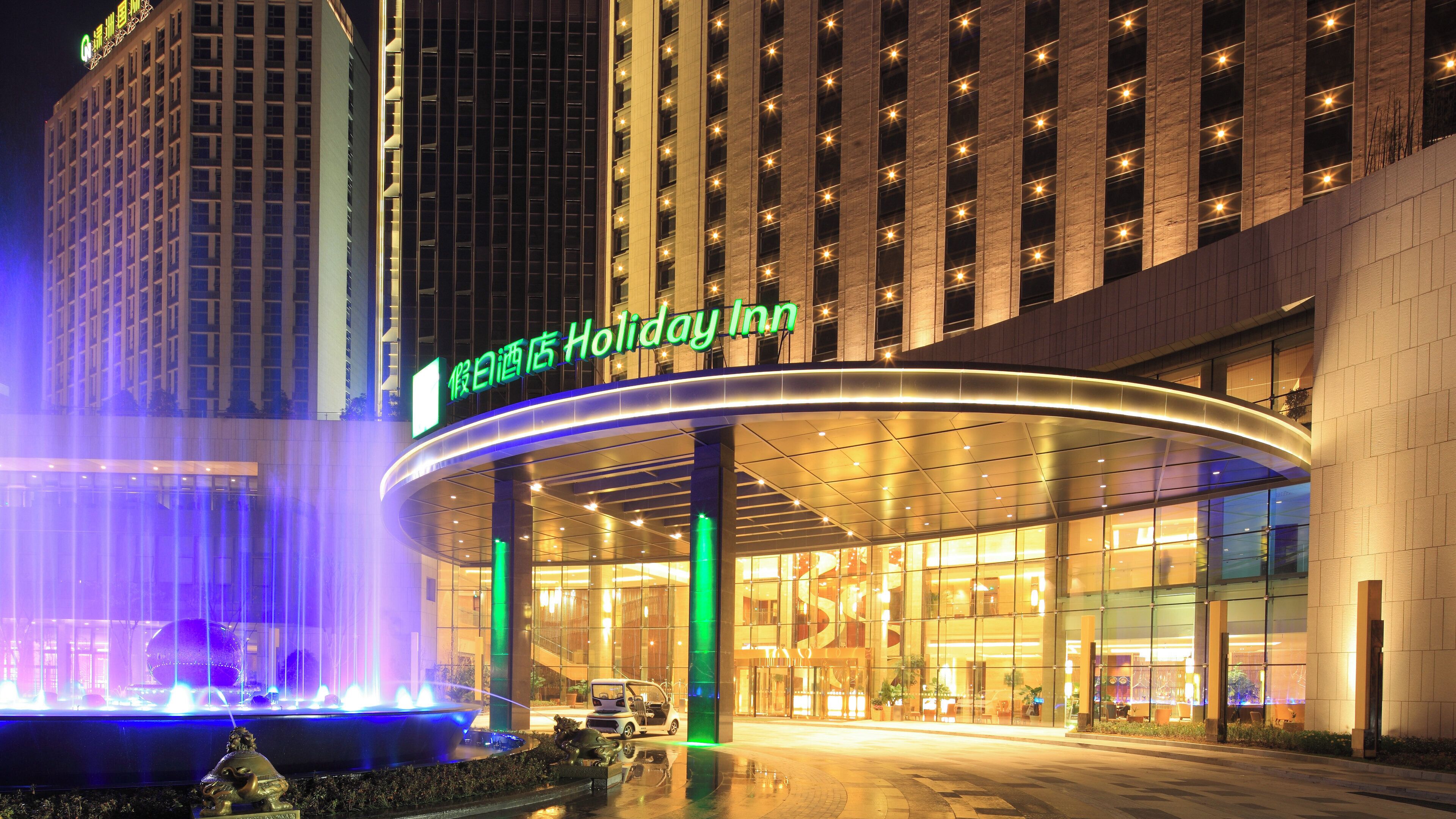 Entrada de un hotel con una fuente, un cartel de 'Holiday Inn' y una fachada de edificio moderna.