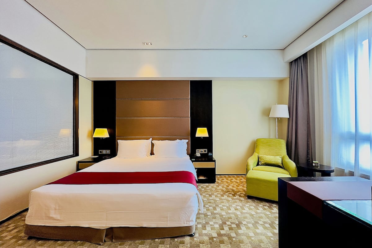 A hotel room with a large bed, two bedside tables with lamps, a green armchair, a desk, and a large window with curtains.