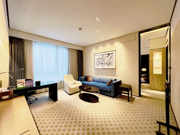 Una habitación de hotel moderna con un sofá azul, un sillón blanco, una mesita de centro, un escritorio con una computadora y un ventanal grande con cortinas.