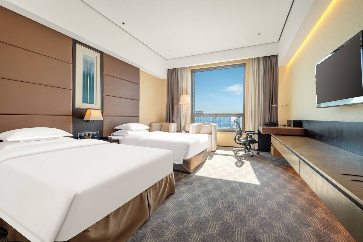 A hotel room with two beds, a desk, a chair, a TV, and a large window with a city view.