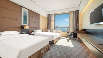 A hotel room with two beds, a desk, a chair, a TV, and a large window with a city view.