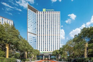 A Holiday Inn hotel with a modern glass facade, surrounded by trees and a well-maintained garden. - Holiday Inn Nantong Oasis Centre by IHG (Nantong)