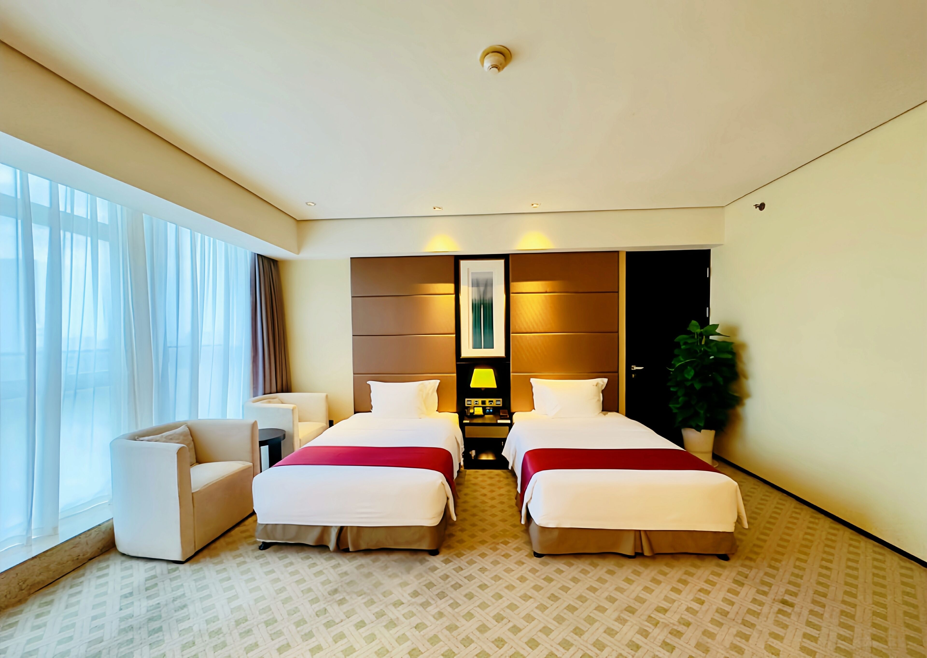 A hotel room with two beds, a desk, a chair, a potted plant, and a large window with curtains.