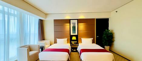 A hotel room with two beds, a desk, a chair, a potted plant, and a large window with curtains.
