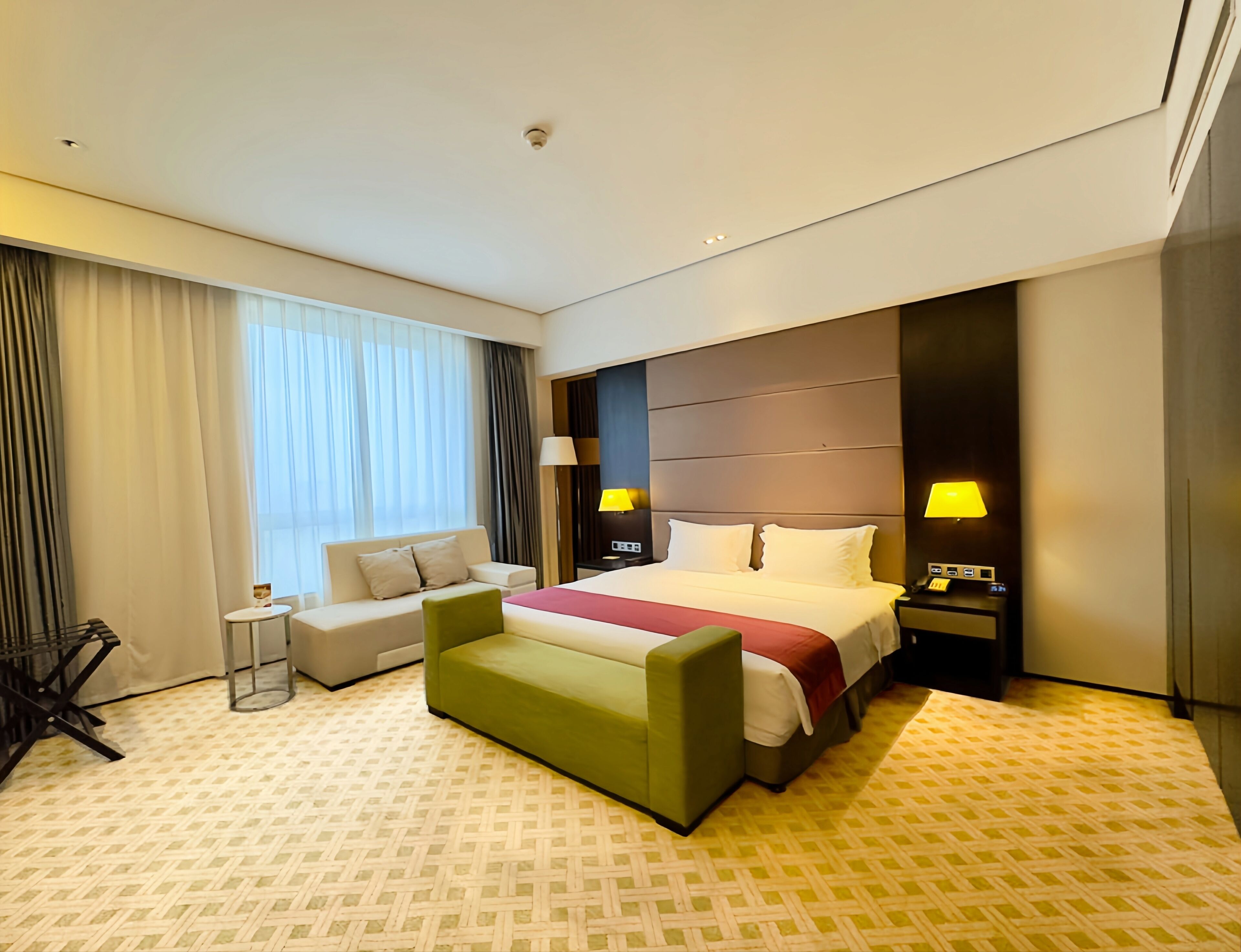 A hotel room with a bed, a green armchair, a small table, and a nightstand with a lamp.
