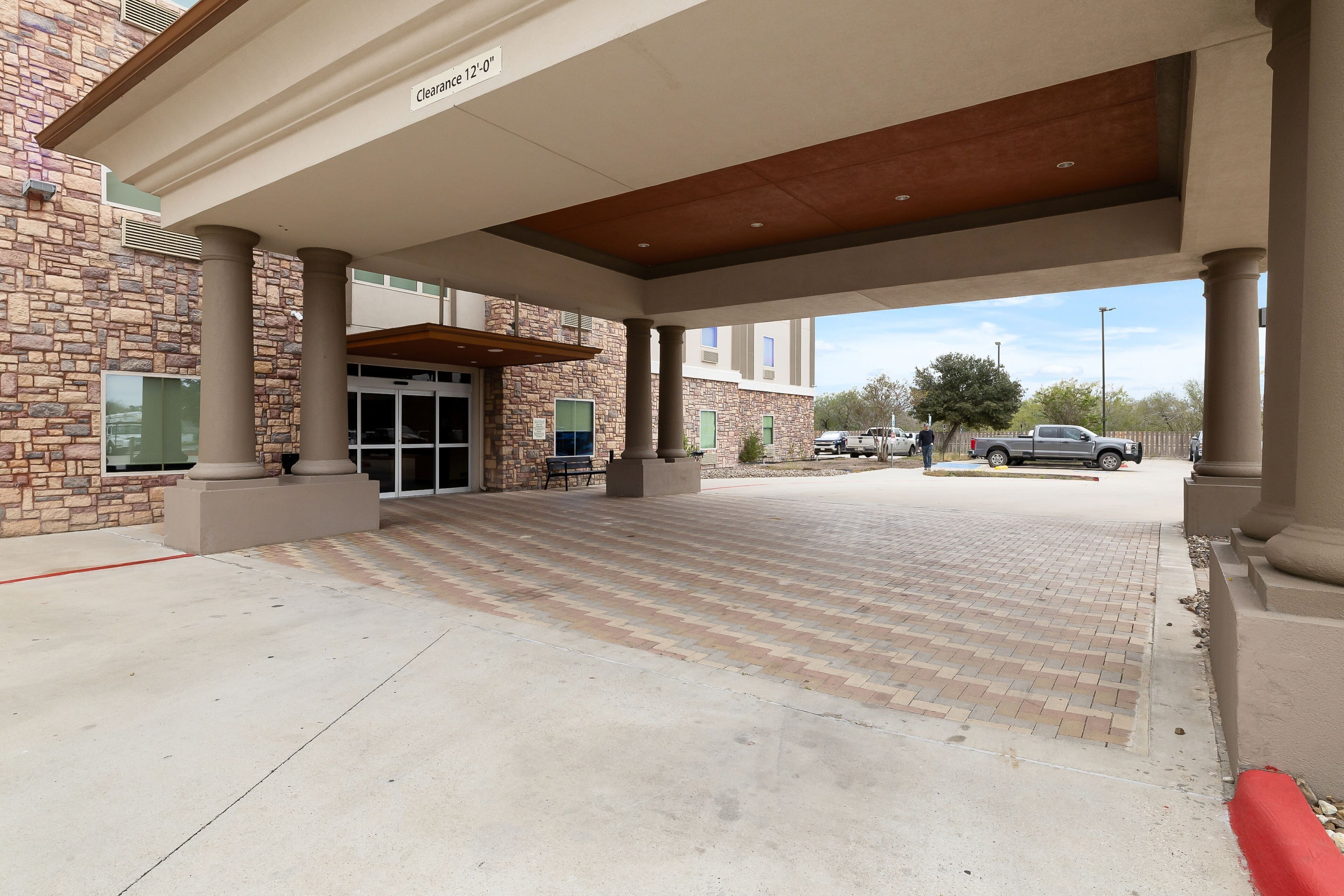 A covered entrance with a brick facade, a clear path leading to the entrance, and a parking area visible in the background.