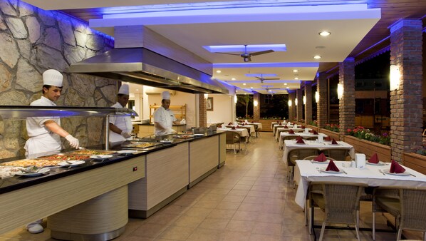 Uma área de buffet com chefs preparando comida, cercada por mesas prontas para refeições.