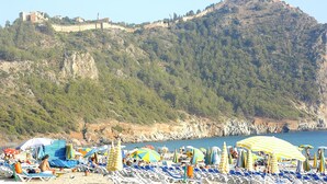 Una playa con filas de tumbonas y sombrillas, un castillo en una colina al fondo y un mar azul cristalino.