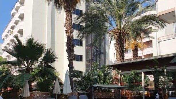 Una piscina con sillas de descanso y sombrillas, rodeada de palmeras y un edificio de varios pisos.