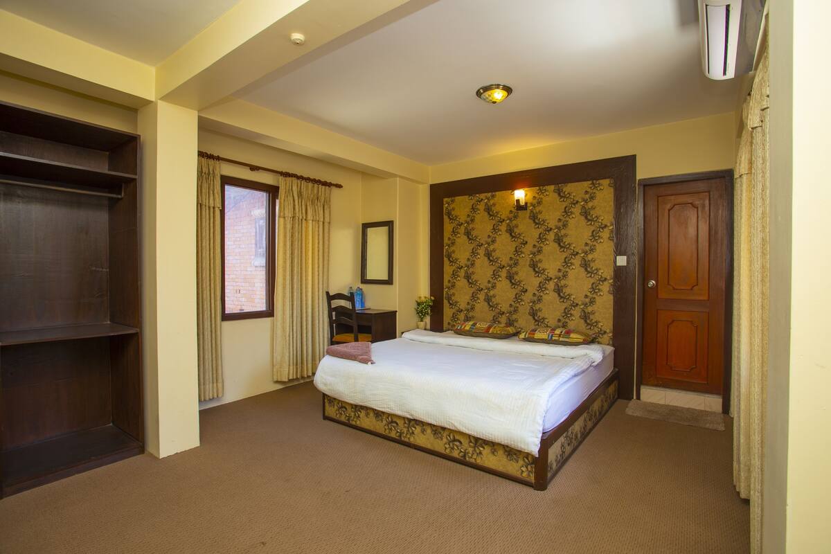 A hotel room with a bed, a desk, a chair, a window with curtains, a door, and an air conditioning unit.