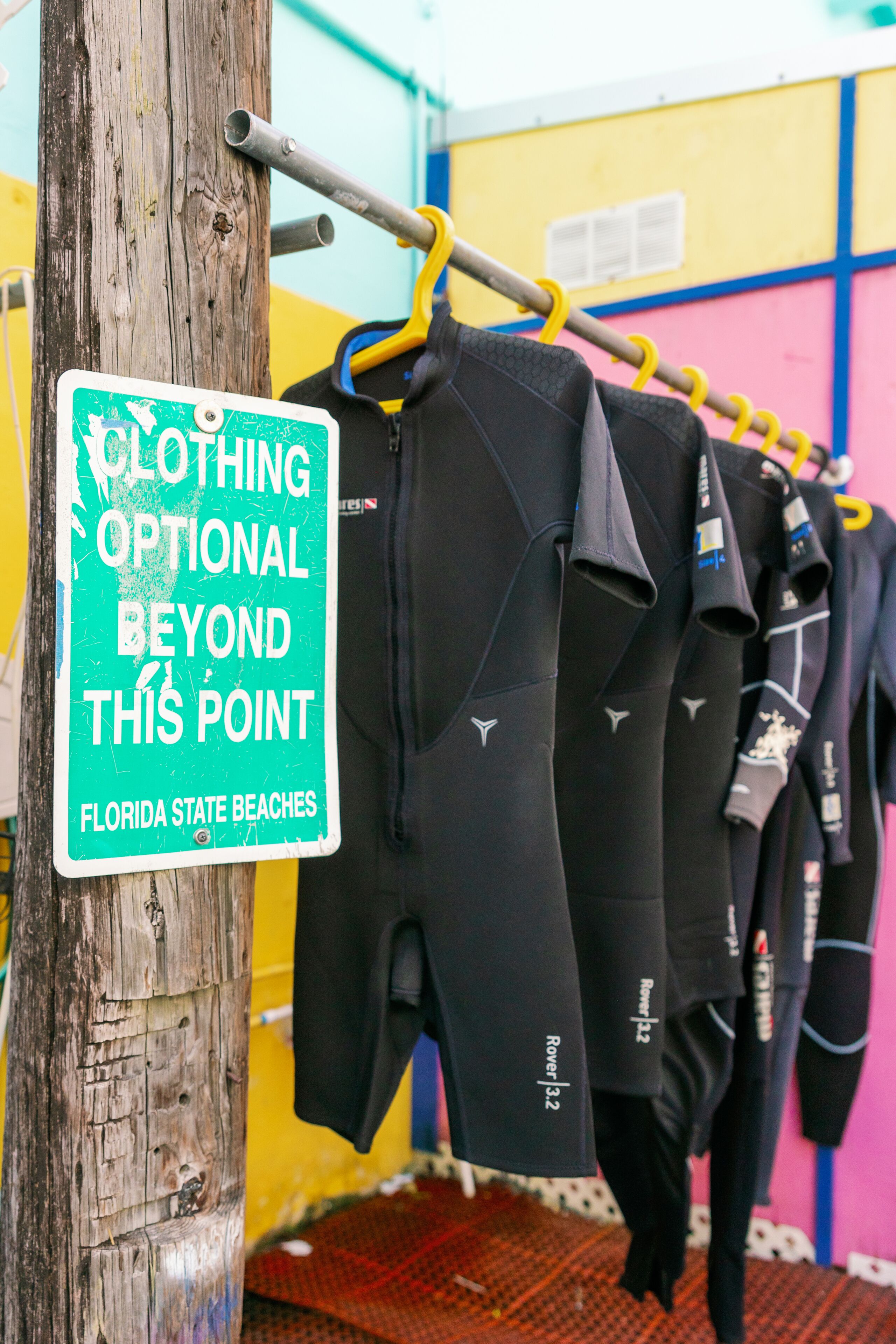 A rack of wetsuits hanging on a wooden post with a sign indicating optional clothing beyond this point.