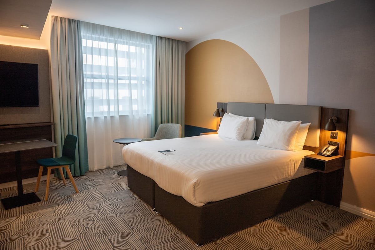 Habitación de hotel con una cama grande, un escritorio, una silla, un televisor y un teléfono sobre la mesita de noche.