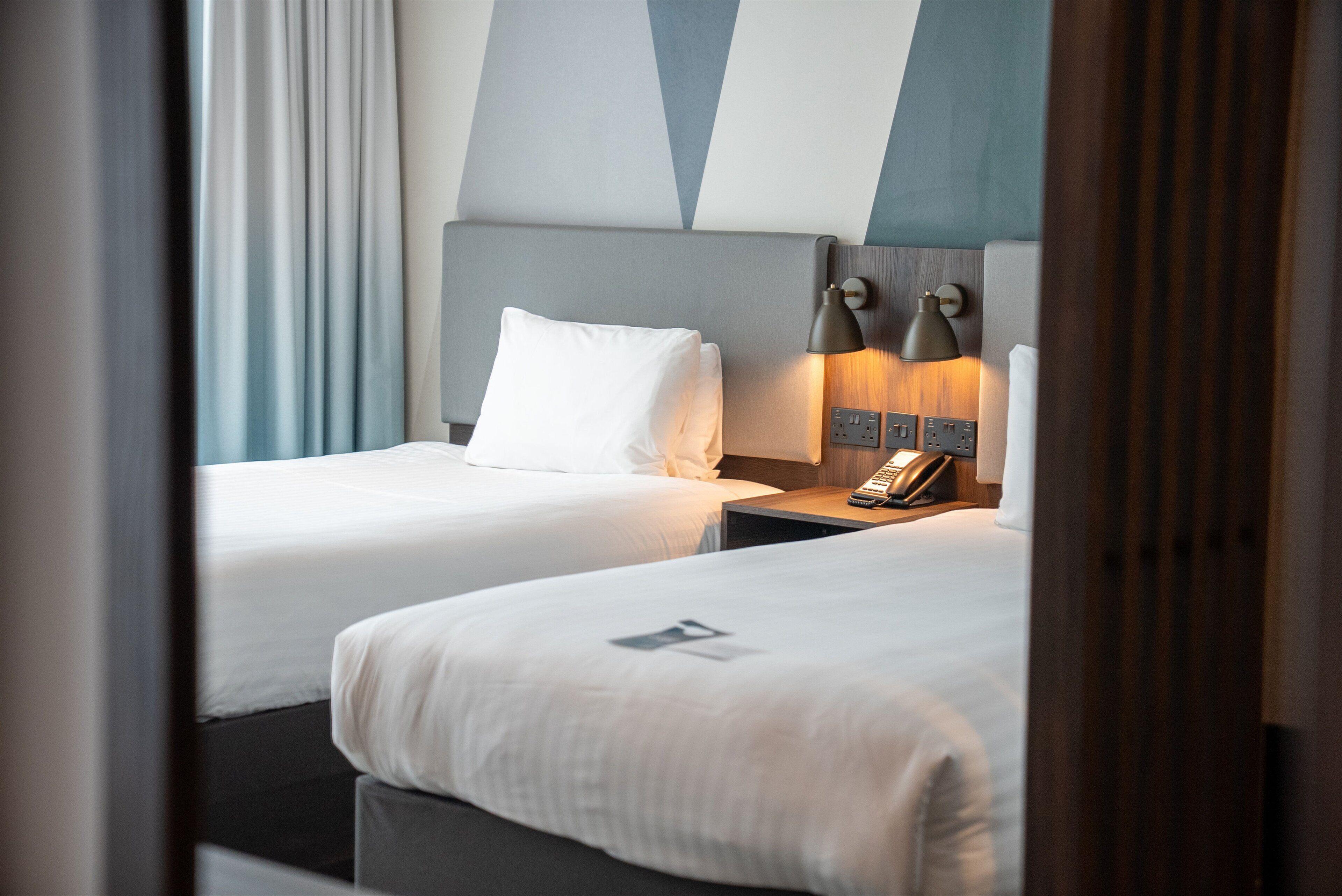 Quarto de hotel com duas camas, uma mesa de cabeceira com abajur e telefone.