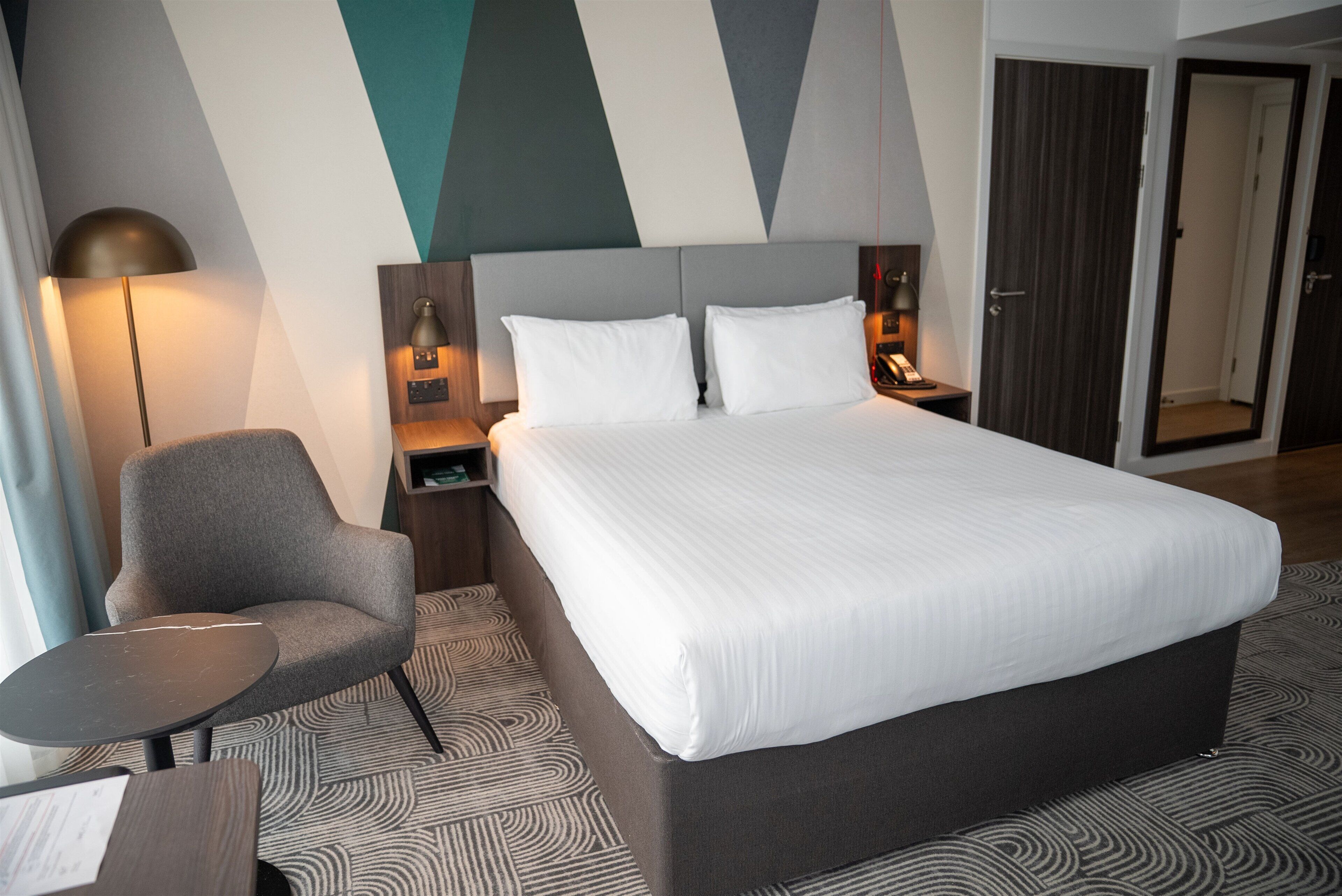 A hotel room with a large bed, a chair, a small table, a lamp, and a wardrobe.