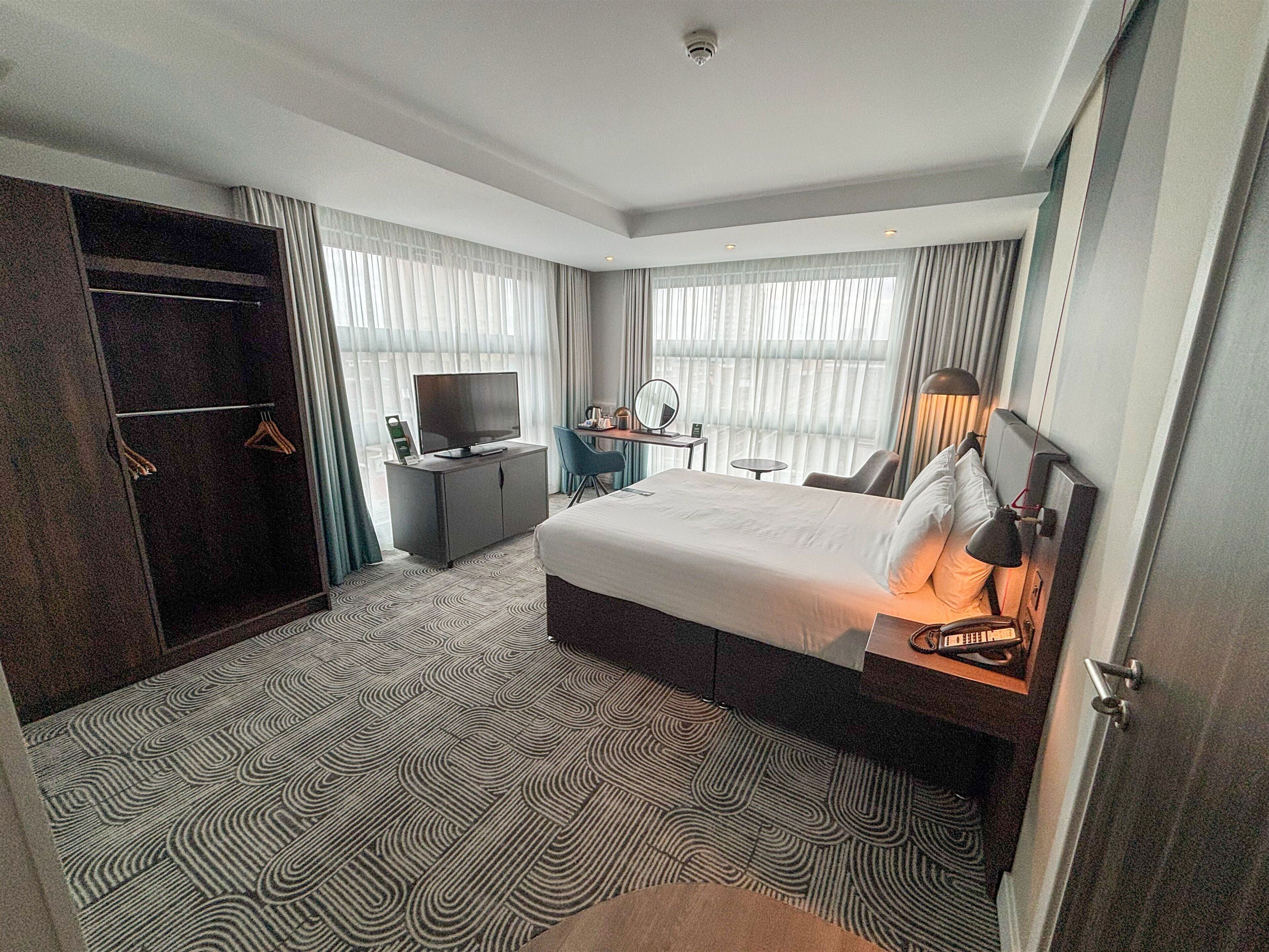 Quarto de hotel com cama, escrivaninha, televisão, guarda-roupa e janela com cortinas.