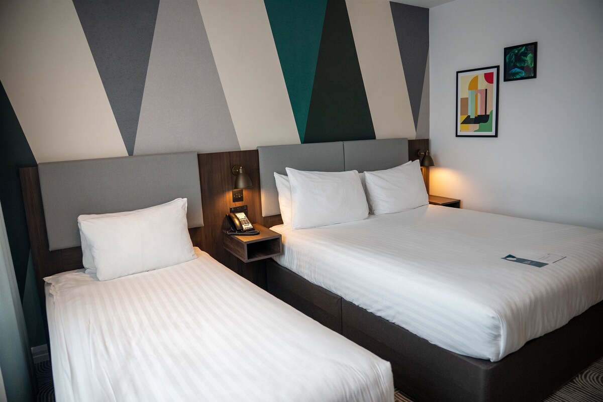 Two adjacent beds with white linens, a nightstand with a telephone, and a wall with geometric patterns and two framed artworks.