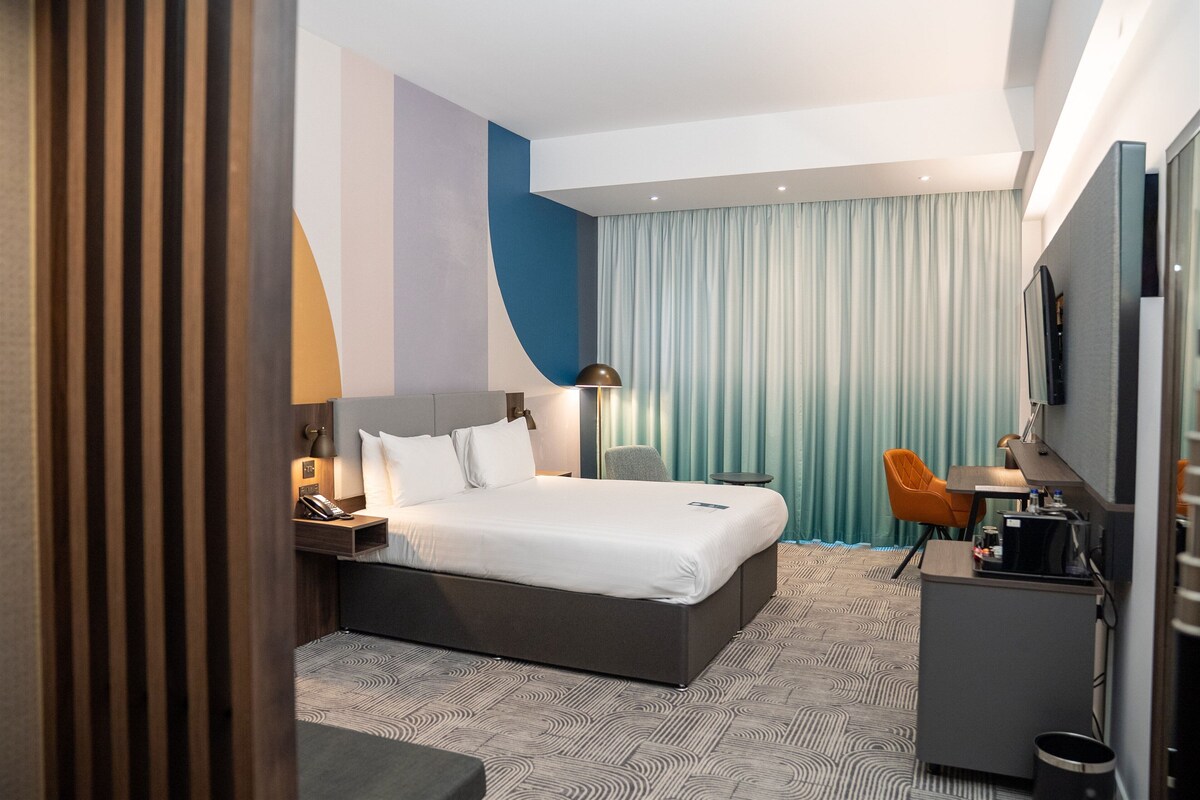 A hotel room with a bed, desk, chair, TV, and curtains.