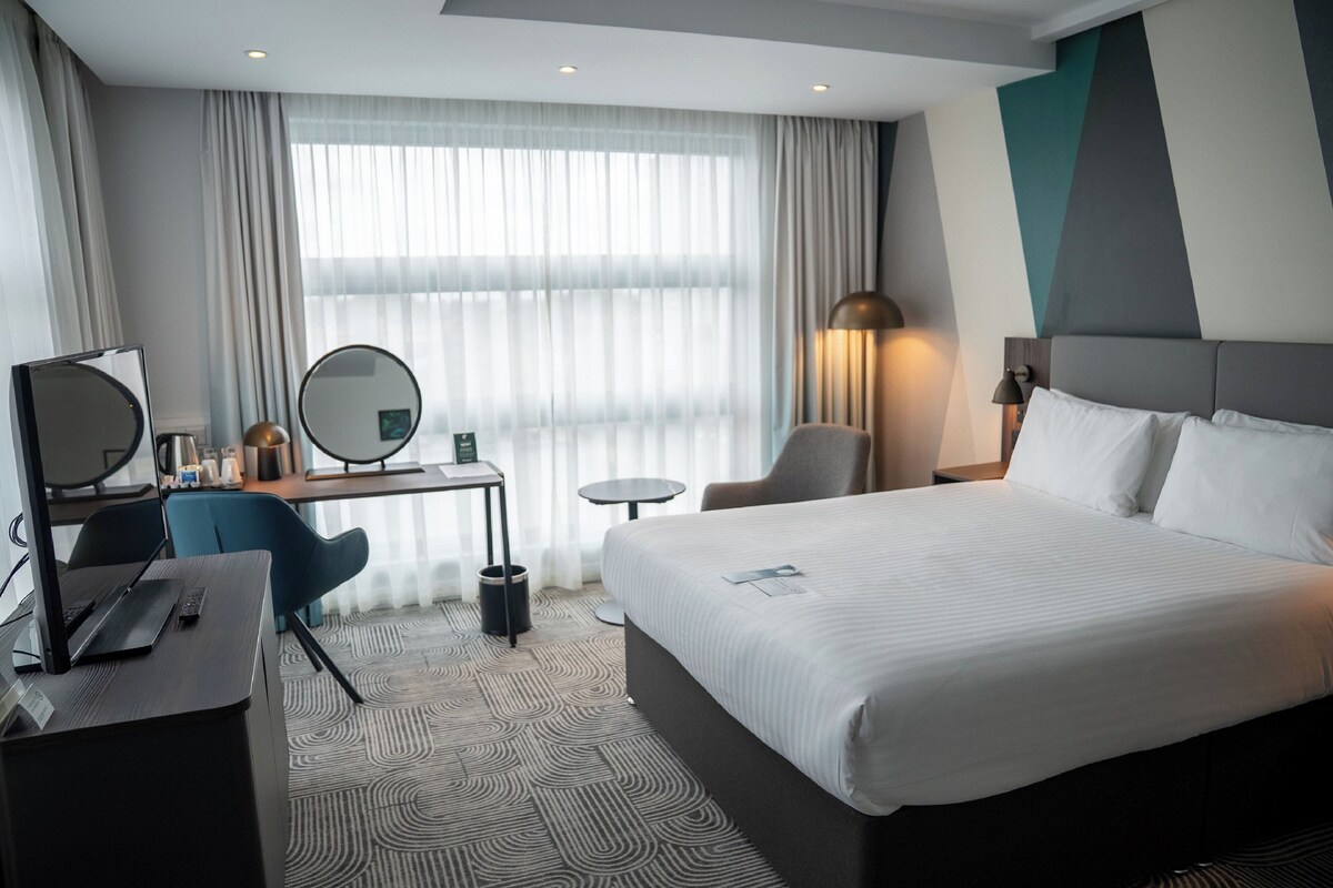 A hotel room with a large bed, a desk with a computer, a chair, a small table, a lamp, and a mirror.