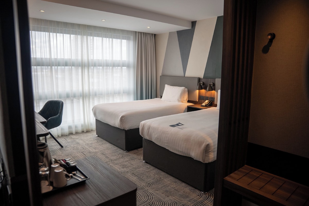 A hotel room with two beds, a desk with a tray of items, a chair, and a large window with curtains.