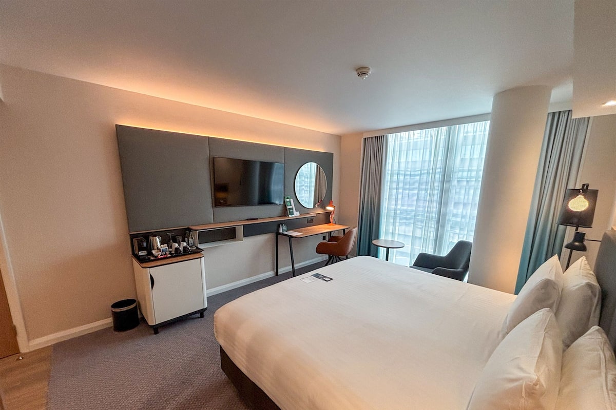 Una habitación de hotel con cama, escritorio, silla, televisor y ventana con cortinas.