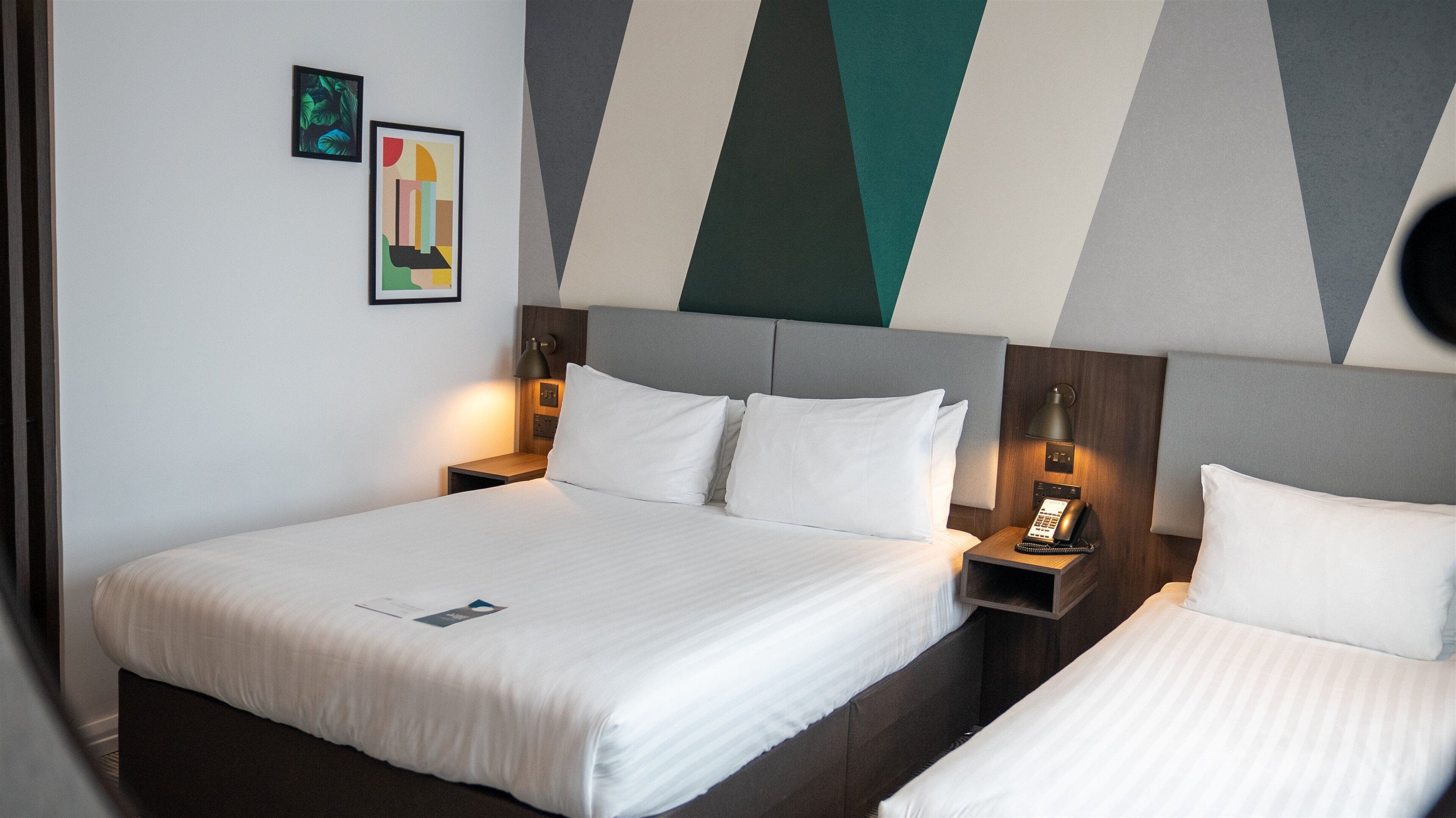 Quarto de hotel com uma cama grande, mesas de cabeceira, um telefone e duas obras de arte emolduradas na parede.