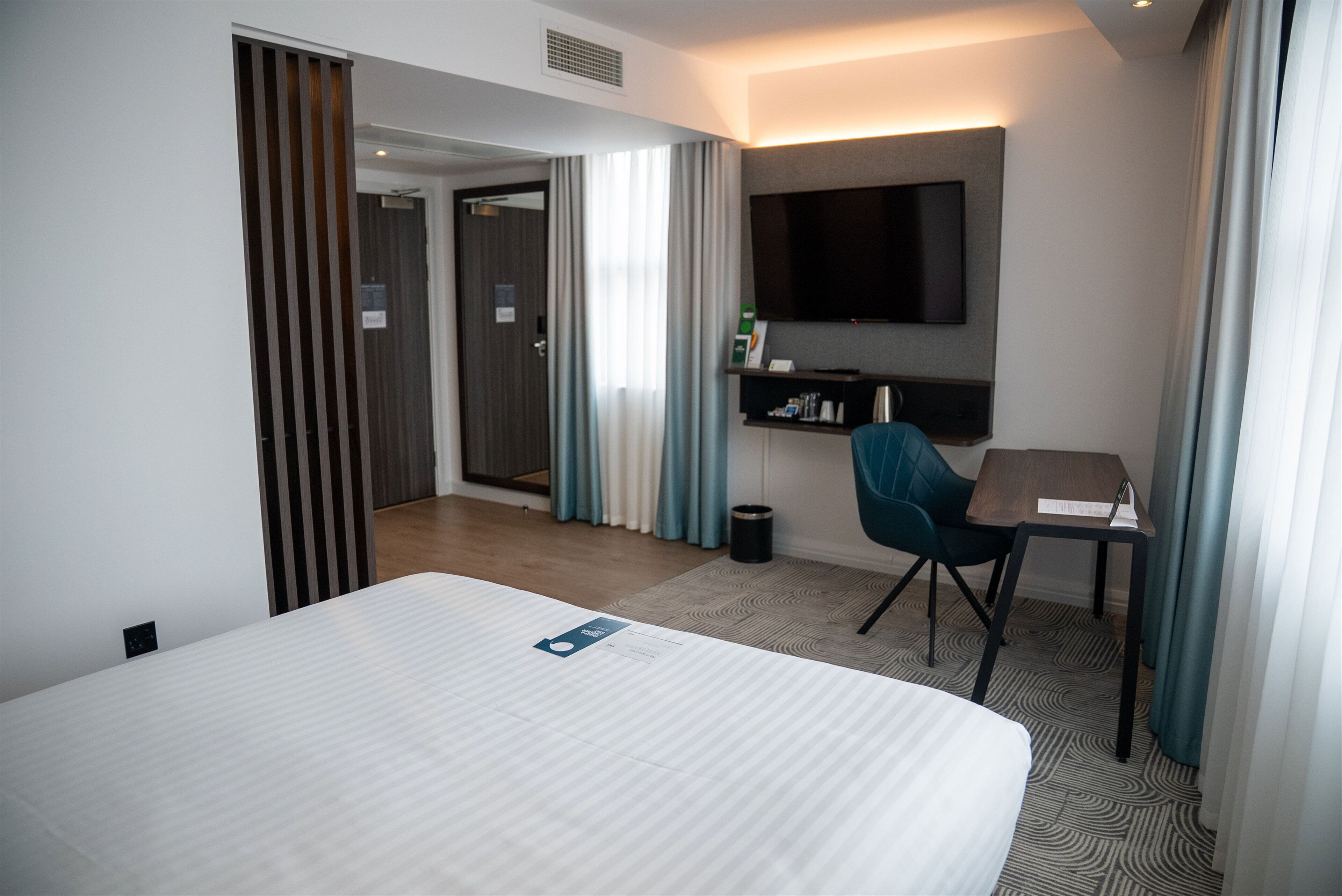 A hotel room with a large bed, a desk, a chair, a TV mounted on the wall, and a door leading to another room.