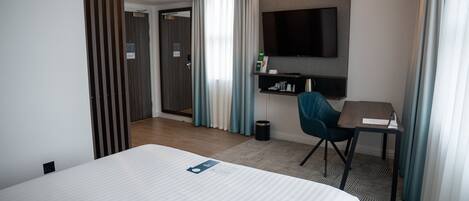 A hotel room with a large bed, a desk, a chair, a TV mounted on the wall, and a door leading to another room.