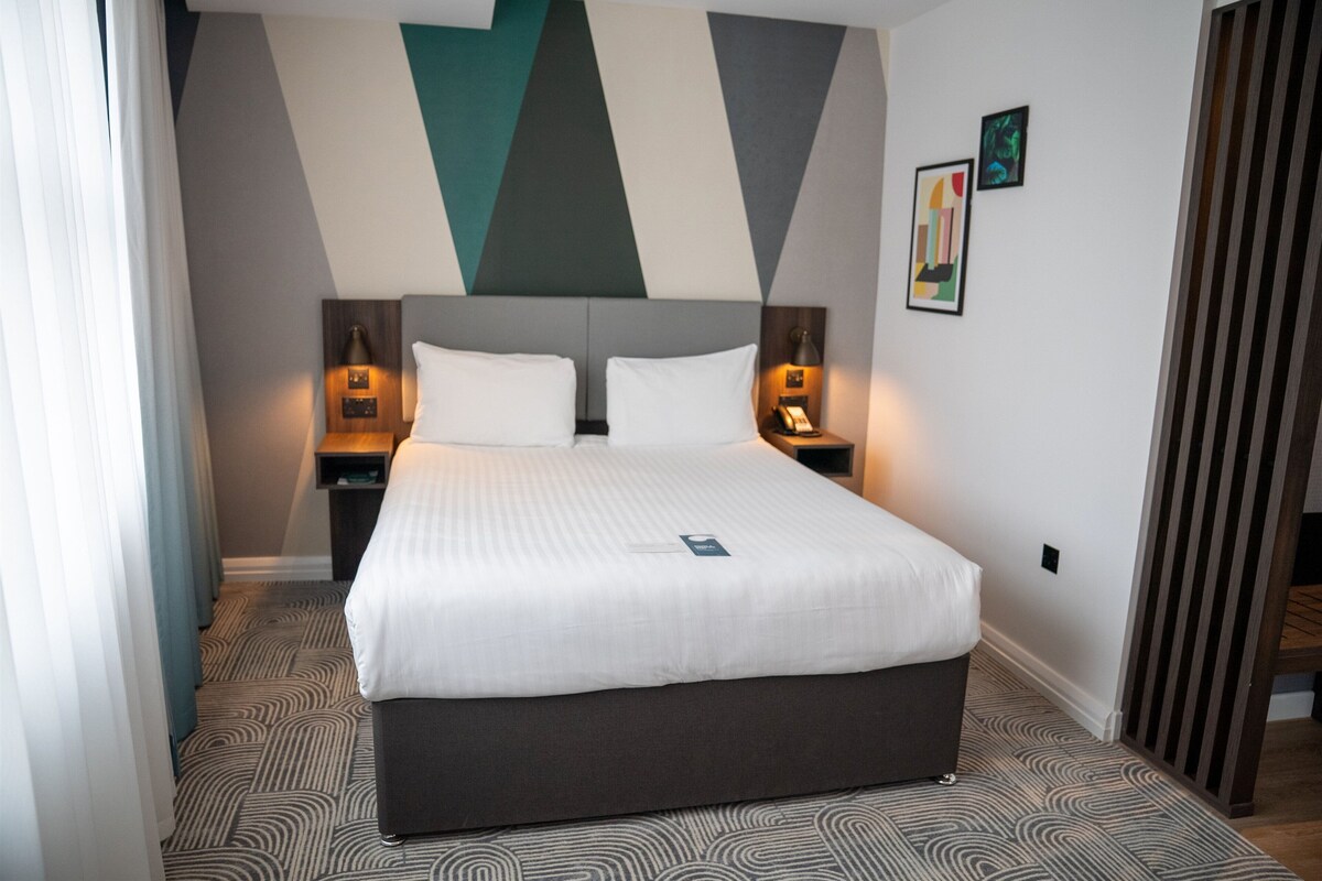 A hotel room with a large bed, two bedside tables with lamps, a headboard, and a patterned carpet.