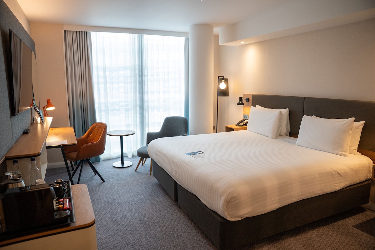 A hotel room with a large bed, a desk, a chair, a small table, and a lamp.