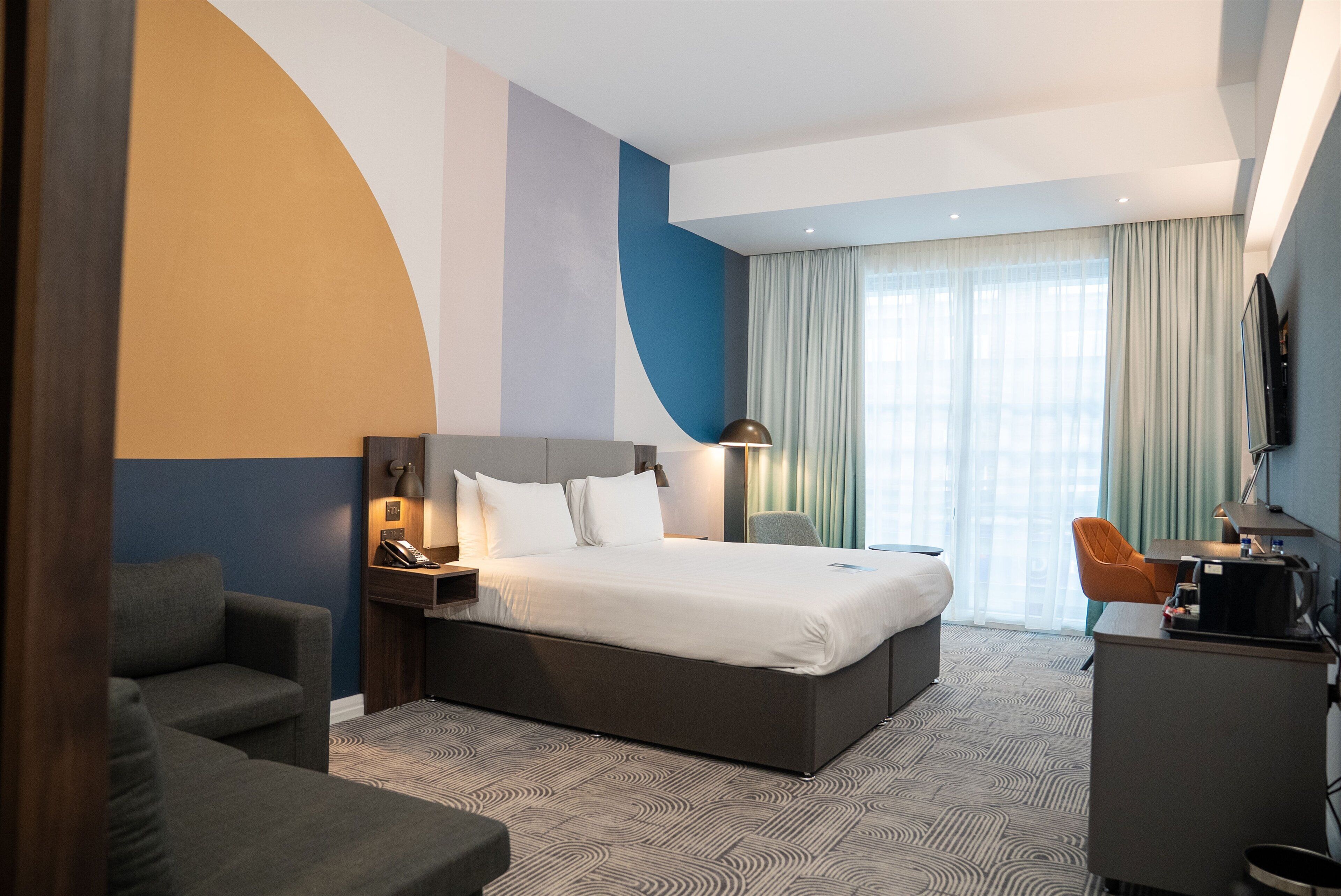 A modern hotel room with a large bed, a desk, a chair, and a TV.