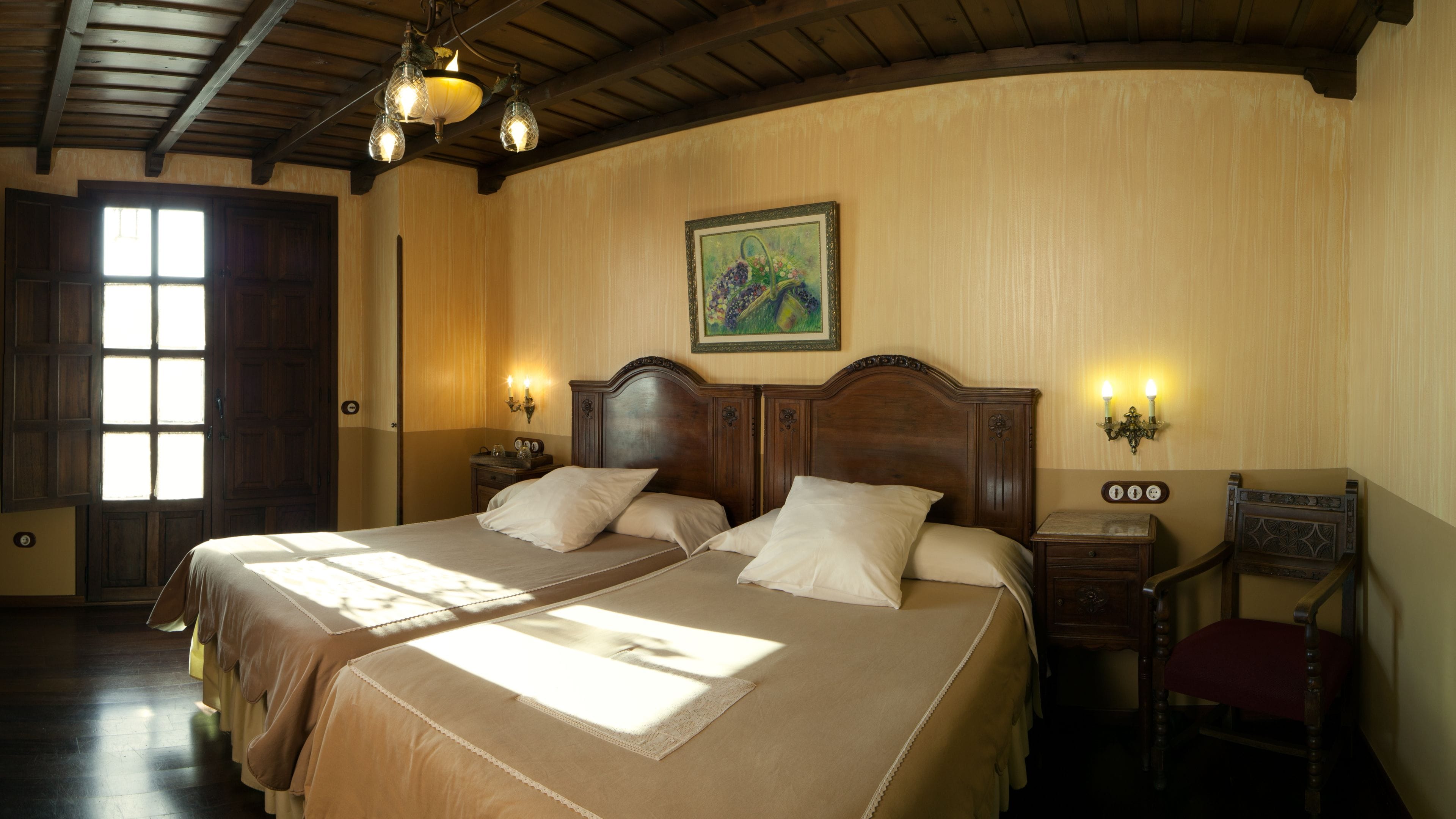 Una cama doble con cabecera de madera, dos mesitas de noche con lámparas, un cuadro en la pared y un ventanal con cortinas.
