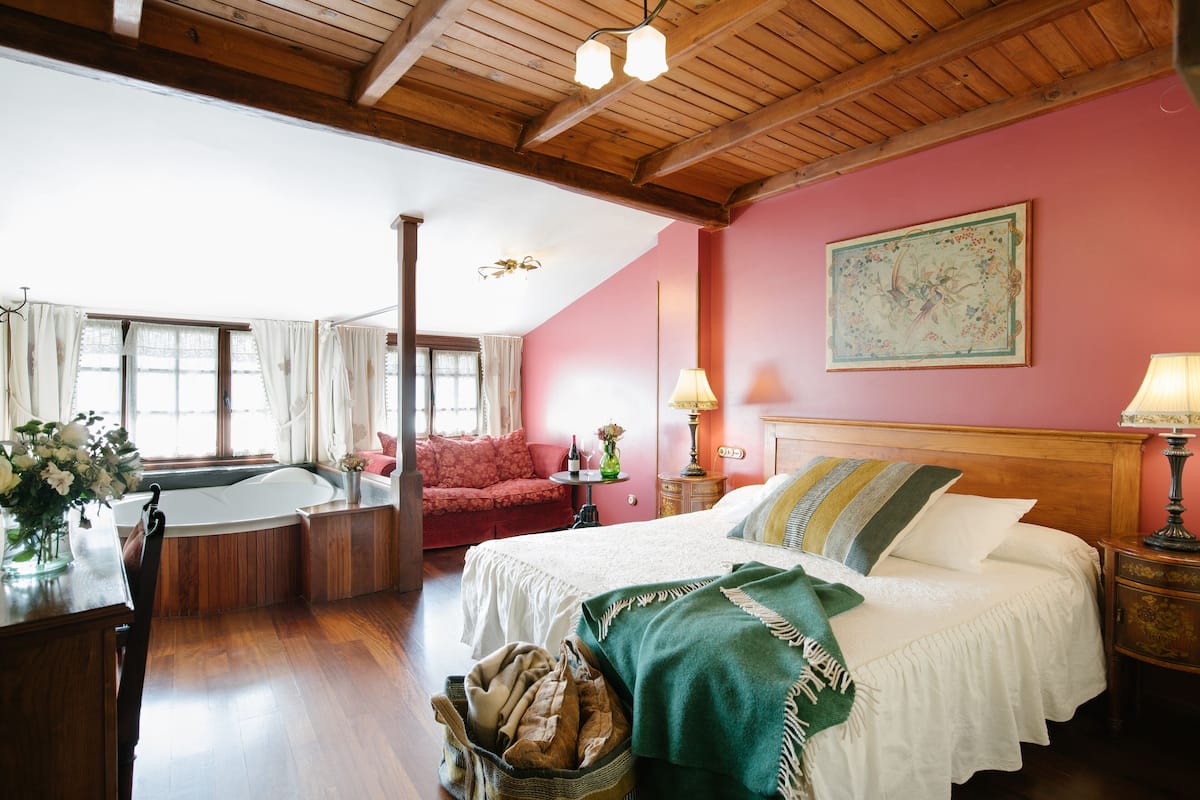 Ein Zimmer mit Holzboden, einem Bett mit grüner und weißer Decke, einem roten Sofa, einem kleinen Tisch mit Lampe und einem großen Fenster mit Vorhängen.