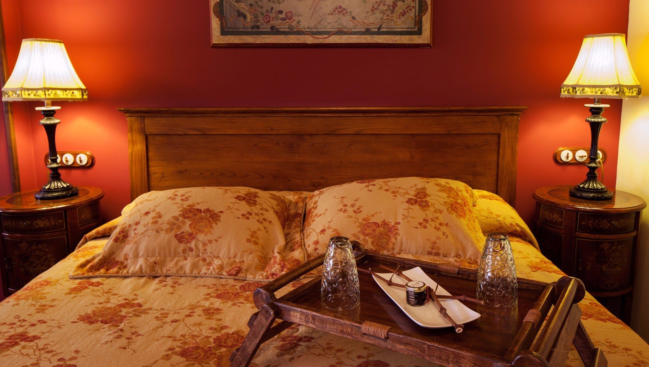 A hotel room with a wooden headboard, floral bedspread, two bedside tables with lamps, and a tray with a bottle and a jar.