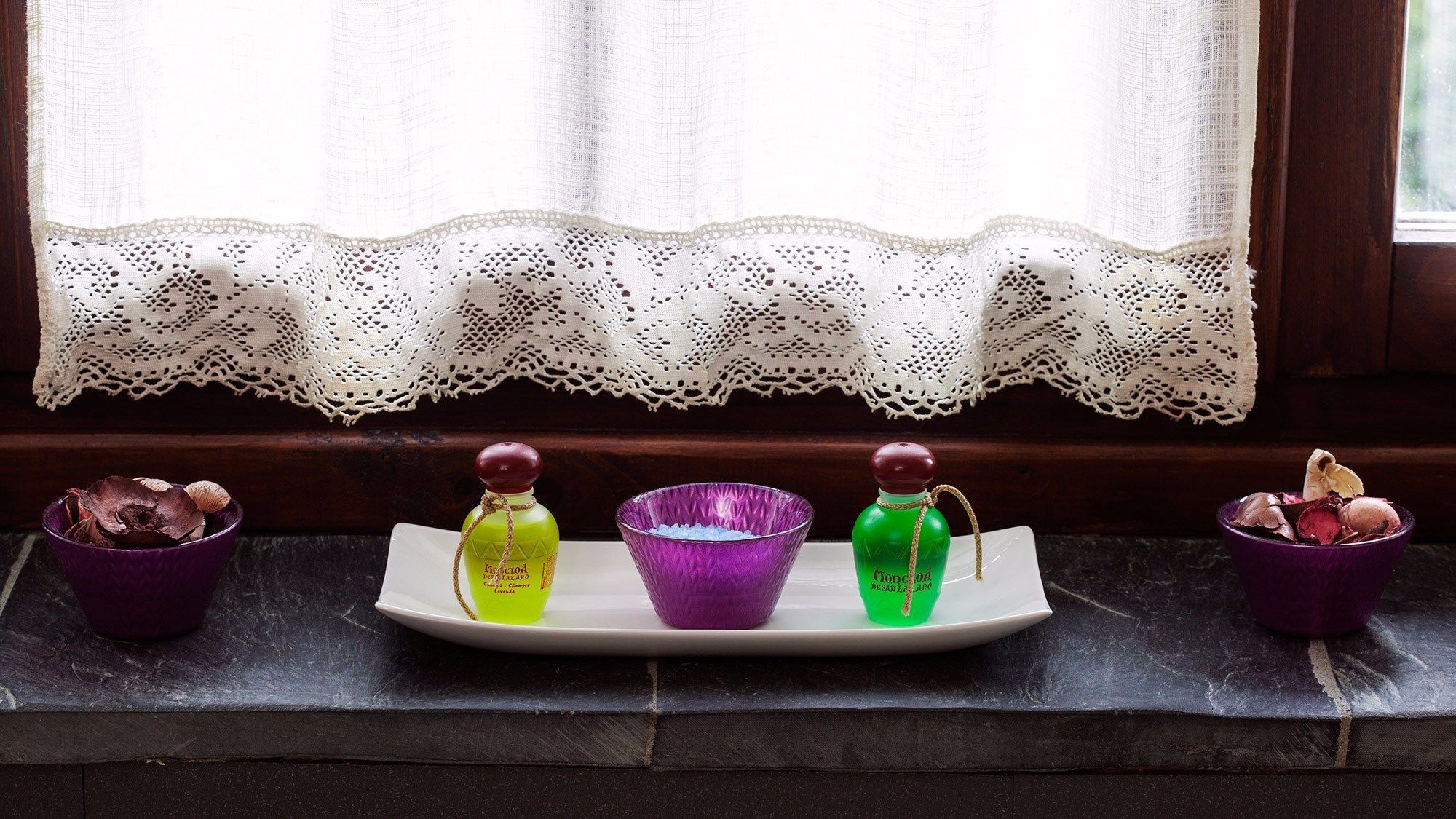 Uma janela com cortina de renda, um parapeito de madeira e dois vasos roxos com flores secas em cada lado.