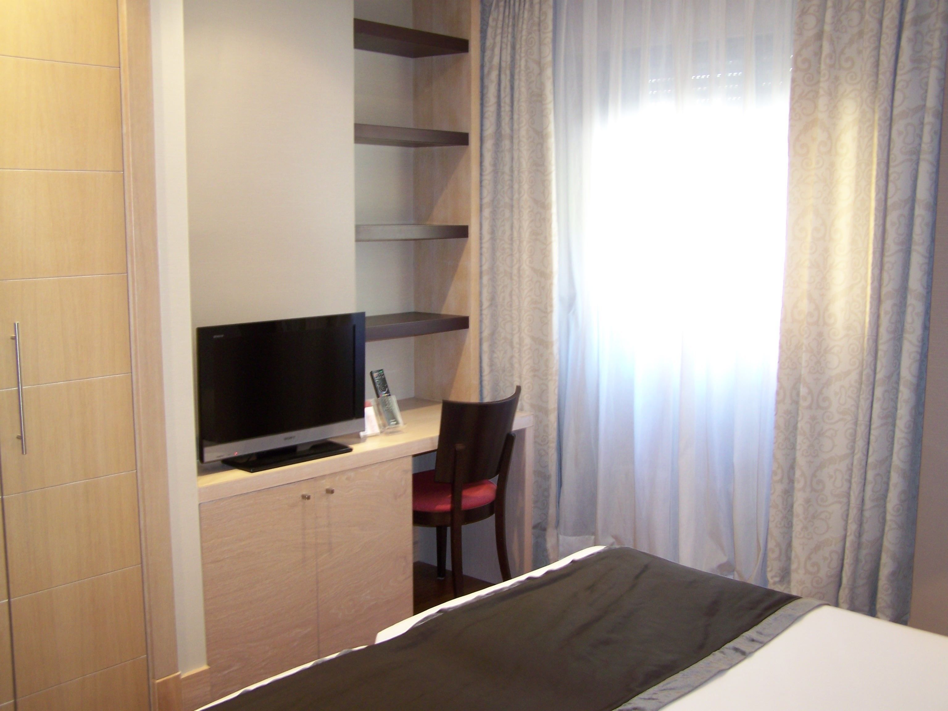 A hotel room with a bed, a TV on a wooden cabinet, a desk with a chair, and a window with curtains.