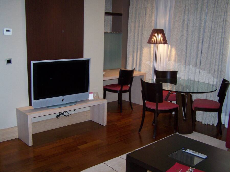 Uma sala de estar moderna com uma televisão de tela plana em um suporte de madeira, uma mesa de jantar com quatro cadeiras e um abajur.