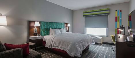 Habitación de hotel con cama, escritorio, silla, mesita de noche, lámpara, ventana con vistas y un cuadro de colores en la pared.