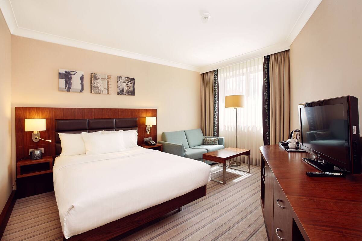 A hotel room with a large bed, a sofa, a TV, and artwork on the wall.