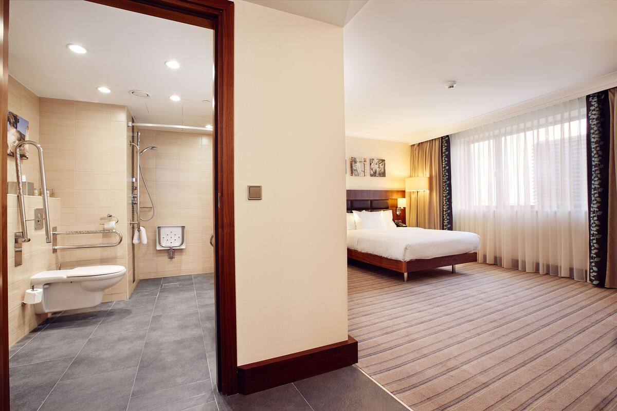 A hotel room with a shower, toilet, bed, and a large window with curtains.