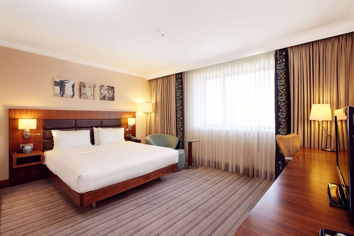 A hotel room with a large bed, bedside lamps, a desk with a chair, and a large window with curtains.