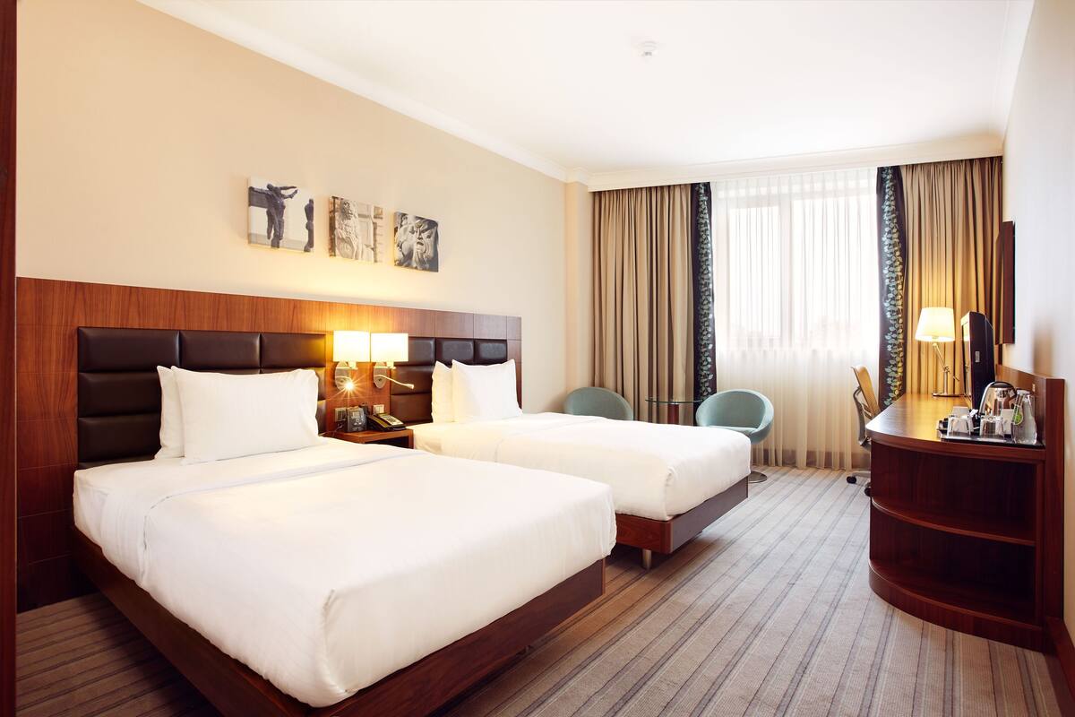 A hotel room with two beds, a desk, a chair, and a large window with curtains.