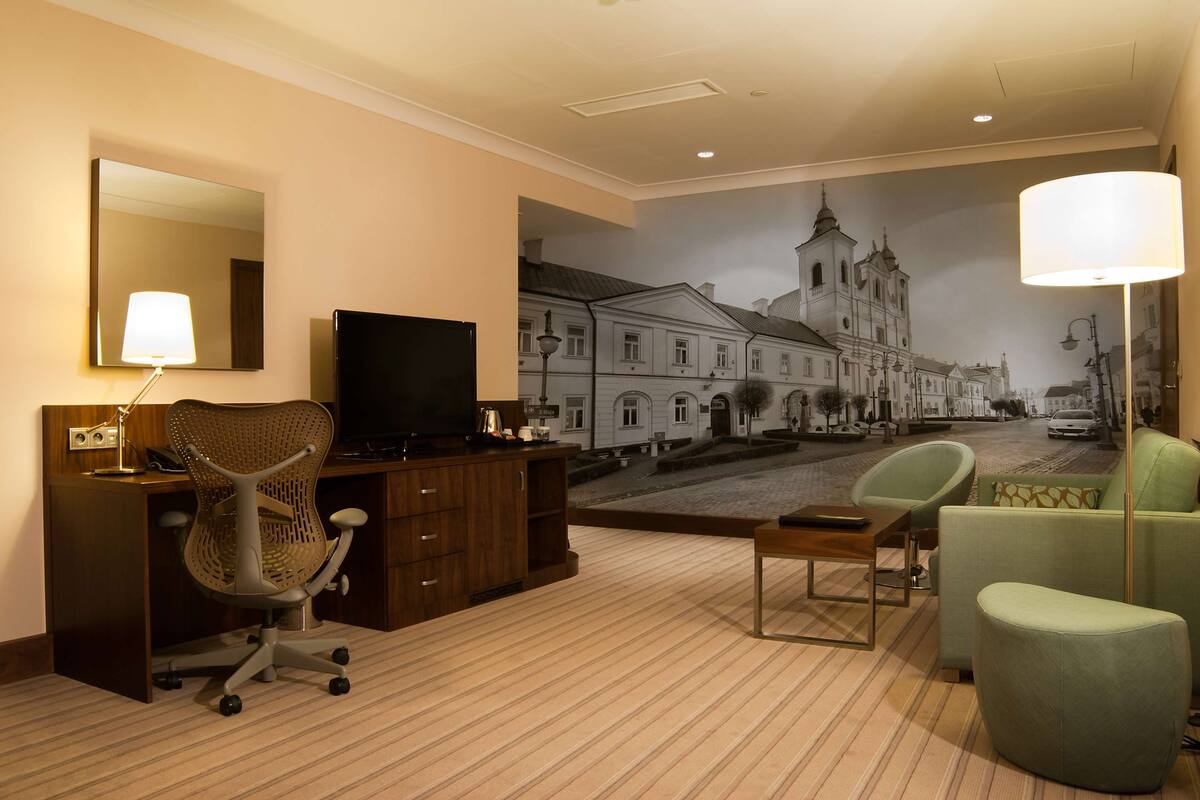 A hotel room with a large mural of a cityscape, a desk with a chair, a television, and a sofa.
