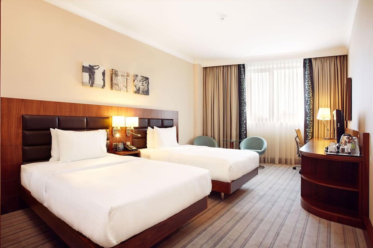 A hotel room with two beds, a desk, a chair, a lamp, and artwork on the wall.