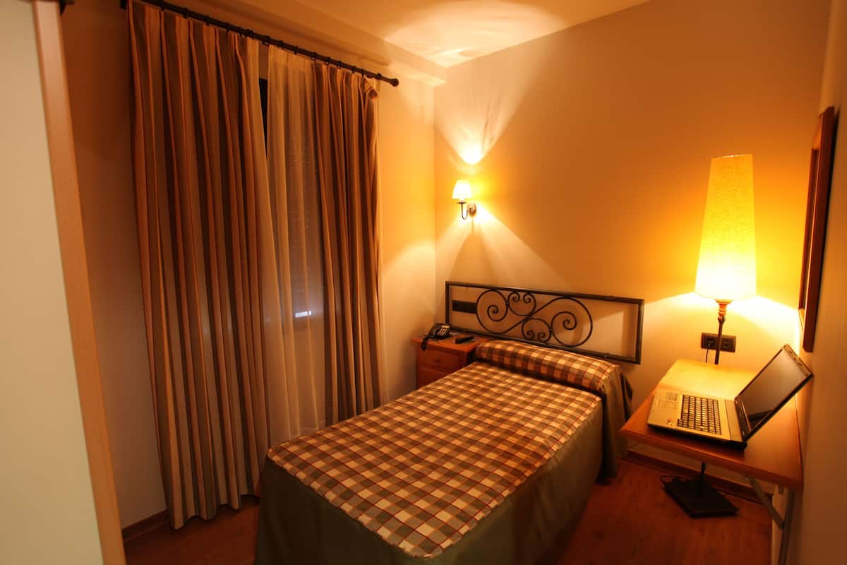 A hotel room with a bed, a nightstand with a lamp, and a laptop on a small table.