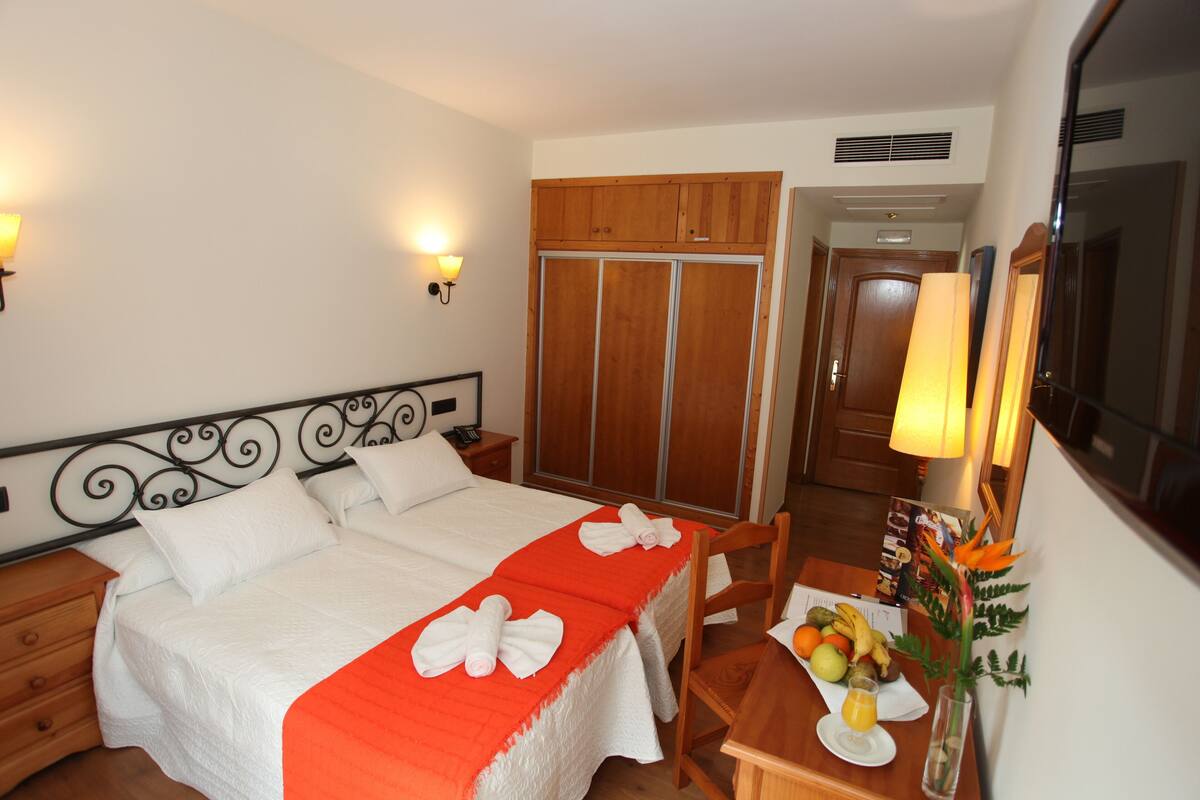 A hotel room with a bed, a wooden wardrobe, a desk with a fruit bowl, and a mirror.