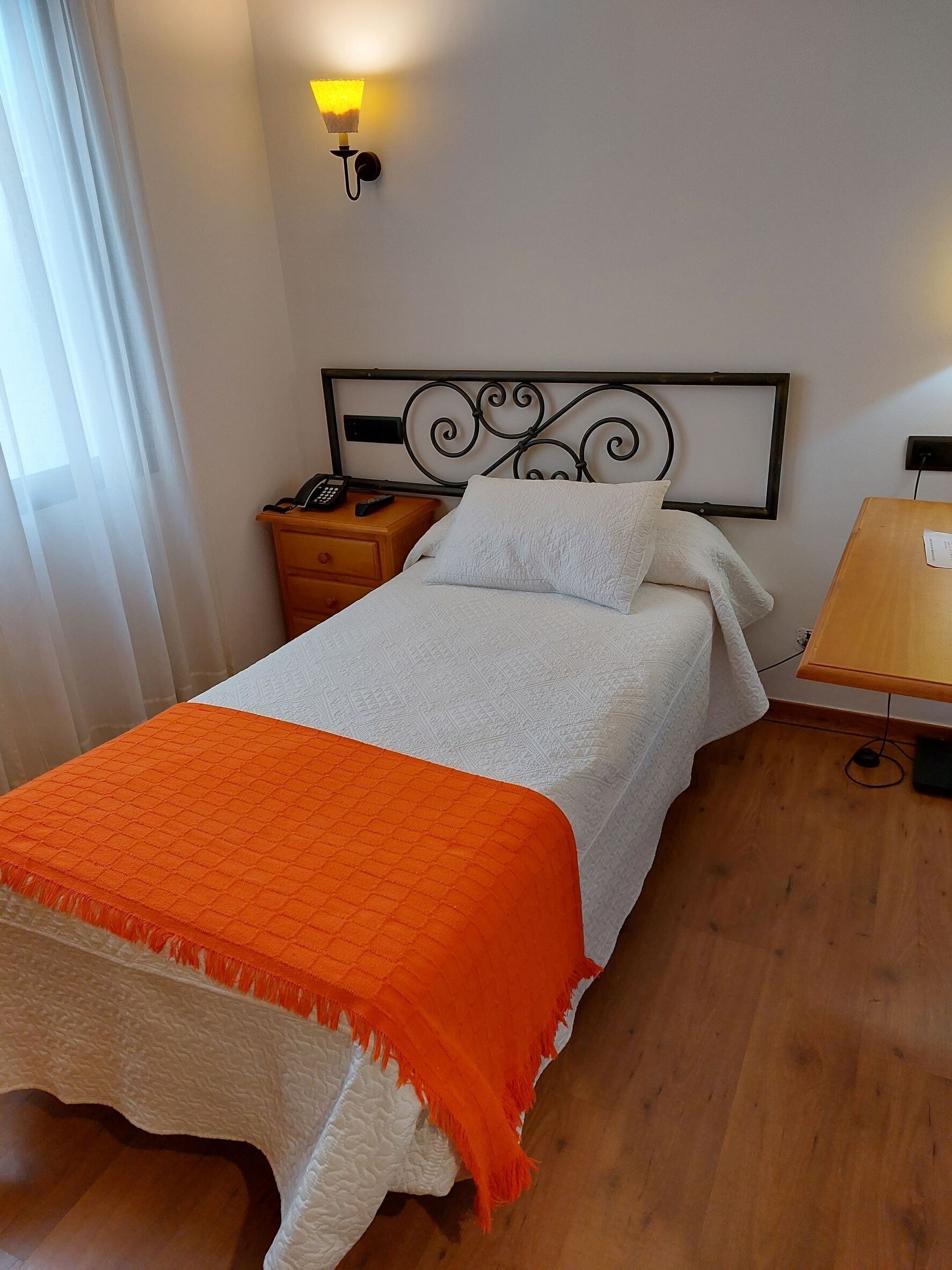 A neatly made bed with a white and orange blanket, a bedside table with a phone and remote control, and a wall-mounted light fixture.