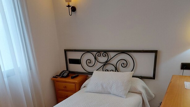 A neatly made bed with a white and orange blanket, a bedside table with a phone and remote control, and a wall-mounted light fixture. - La Bodega Hotel (Ciudad Rodrigo)